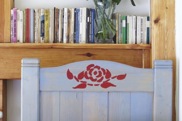 Pojedyncze łóżko z wymalowaną różą na zagłówku. Nad łóżkiem półka z książkami i wazon z różami.